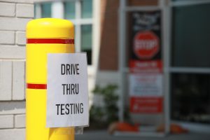 A storefront parking lot with a sign reading Drive Thru Testing taped to a safety bollard.