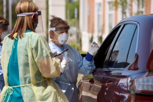Two health care workers in scrubs, plastic protective gear and masks speak with a person through the driver's window of a vehicle. 