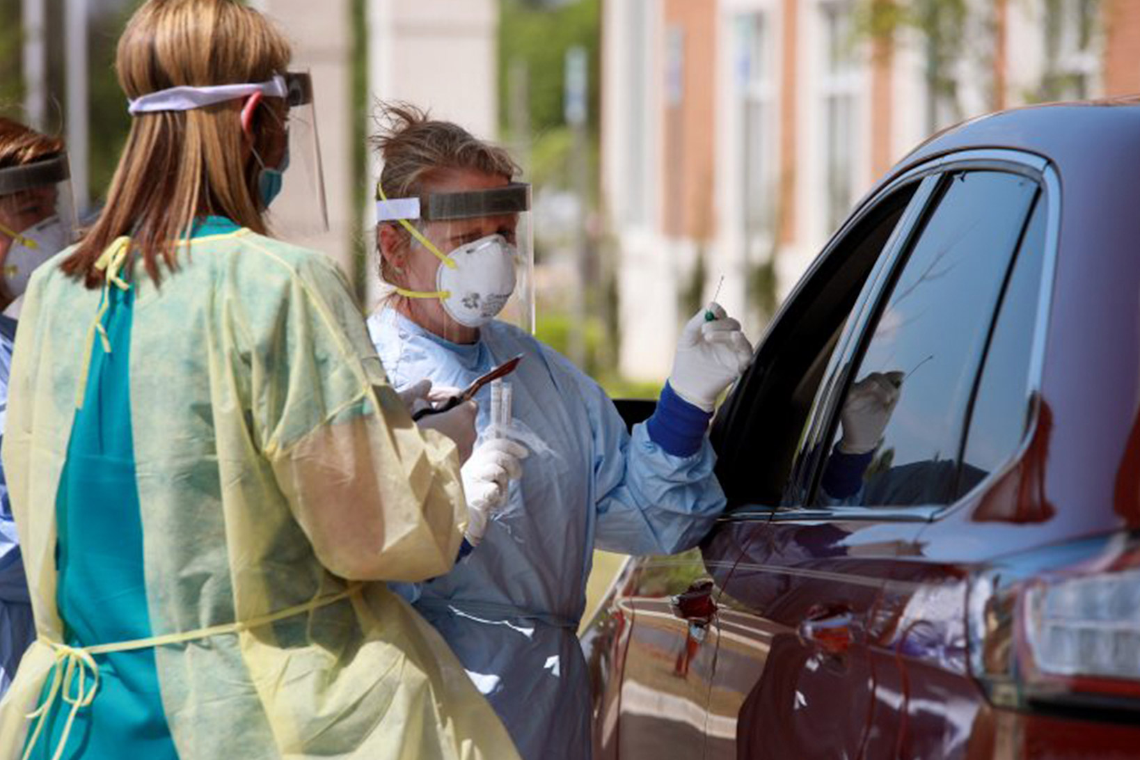 In a parking lot, two Lexington Medical clinicians hold a swab for COVID-19 testing for a patient in a red sedan.