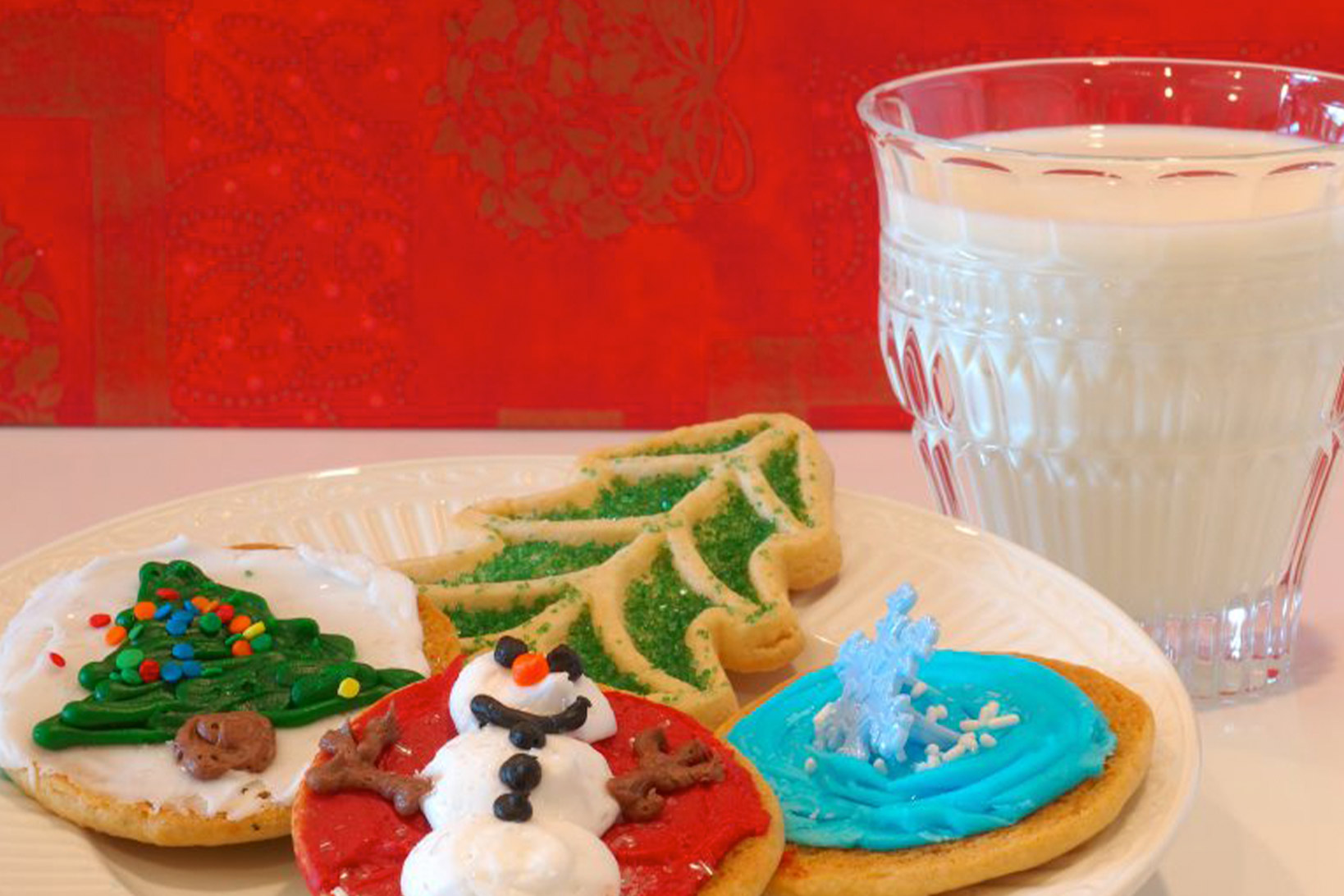 Four holiday-themed cookies on a white plate next to a glass of milk against a red backdrop.