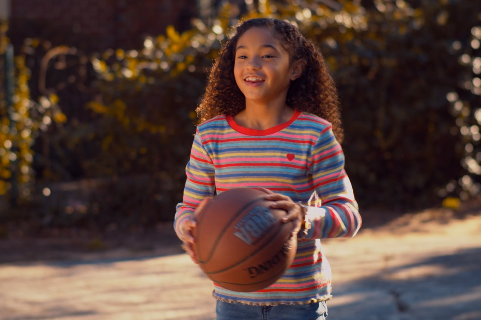 A little girl in a colorful shirt smiling while getting ready to pass a basketball to someone out of view outside on a sunny day.