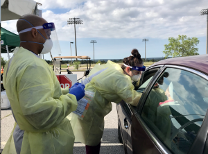 Two health care workers in scrubs, plastic protective gear and masks speak with a person through the driver's window of a vehicle. 