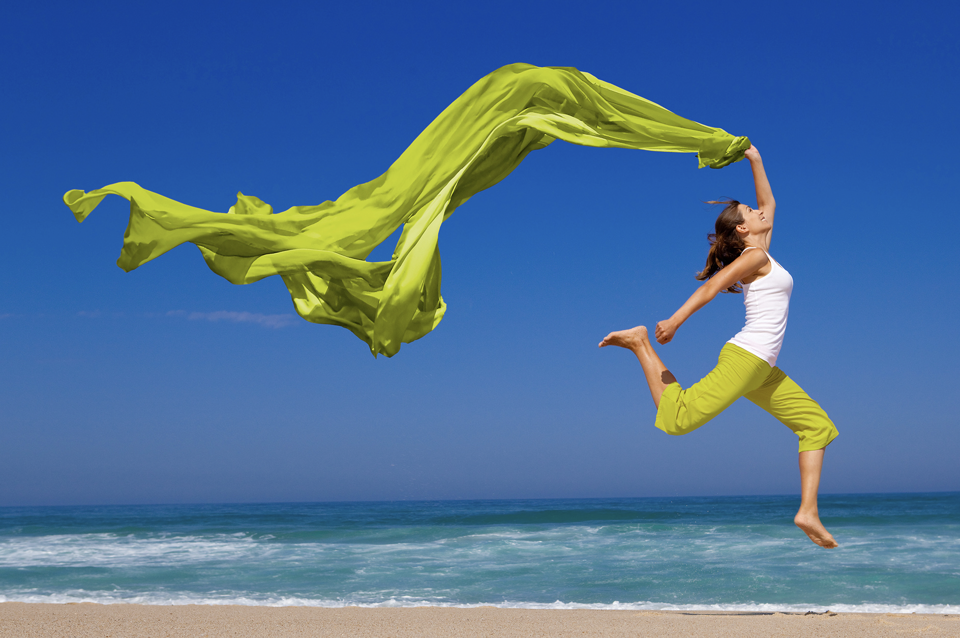 Middle aged woman leaping on beach with green scarf in her hand
