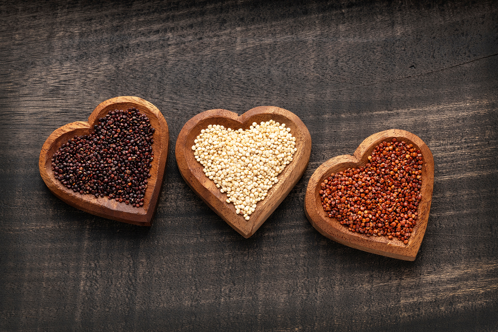 Various colors of quinoa in three heart shaped wooden bowls in black table