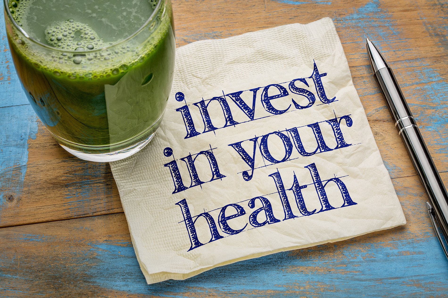 Green smoothie on wood table with napkin that reads "invest in your health'