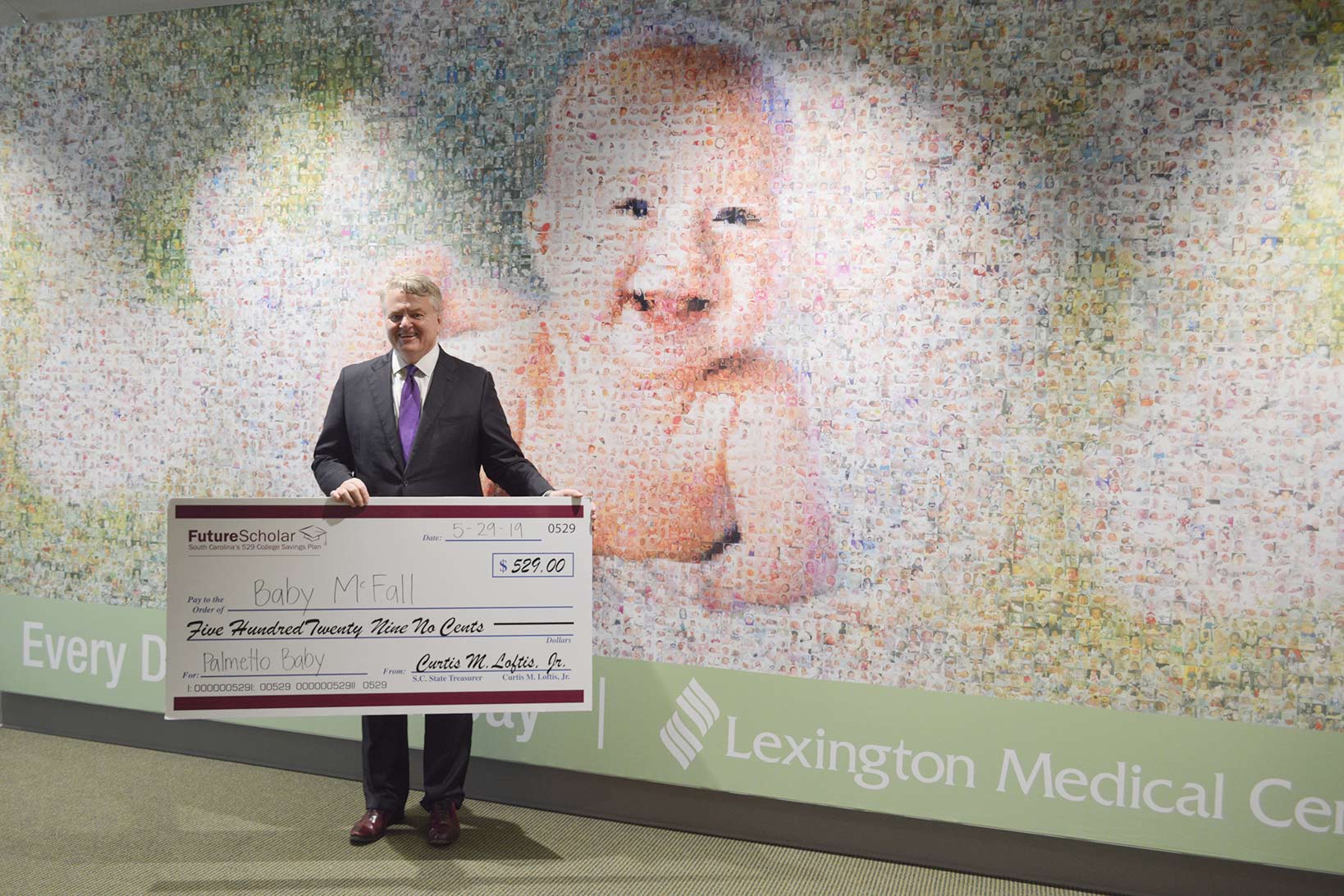 Treasurer Loftis holding a large check addressed to Baby McFall posing in front of the baby mosaic in the new patient care tower.