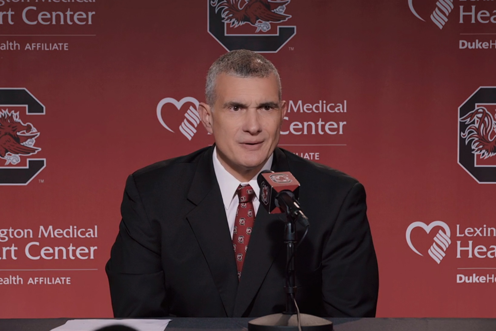 UofSC basketball coach Frank Martin in a suit at a press conference, sitting in front of a garnet microphone against a background with the UofSC logo and the Lexington Medical Heart Center logo.