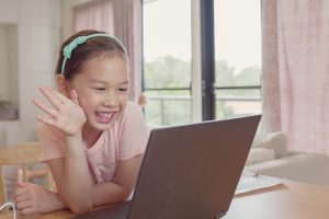 A little girl smiling and waving at a laptop screen.