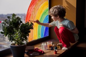 A little boy crouching on a windowsill with a plant on it, intently painting a rainbow on the window.