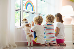 Three children smiling and peering out of a window with a rainbow painted on it while their dog also looks out the window.